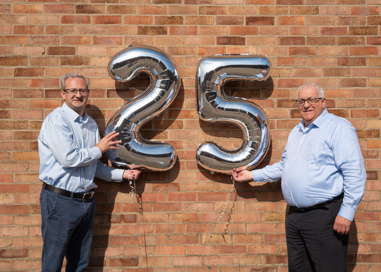 A photo of the two directors holding silver balloons than read '25'. They stand in front of a red brick wall. Both men wear a blue shirt, glasses, and give the camera a smile. They hold the silver balloons in the middle between them.