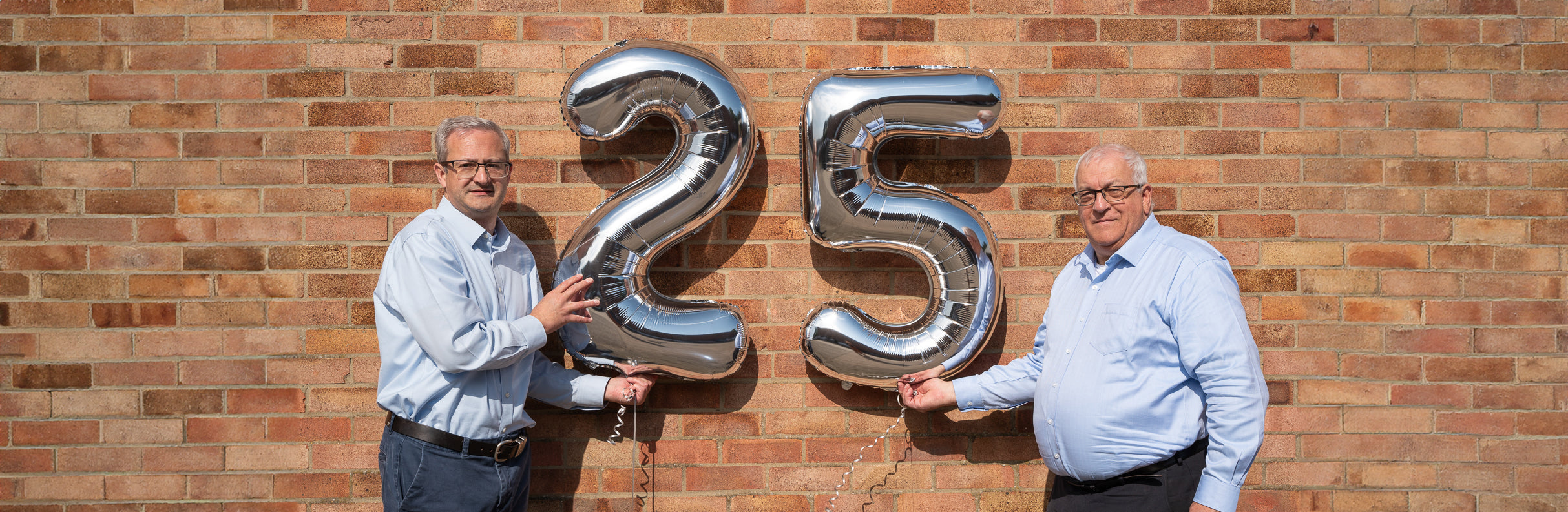25 Balloons A photo of the two directors holding silver balloons than read '25'. They stand in front of a red brick wall. Both men wear a blue shirt, glasses, and give the camera a smile. They hold the silver balloons in the middle between them.
