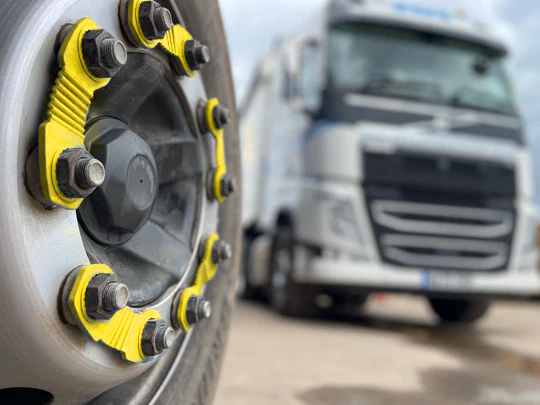 A close up of yellow zafety devices fitted to an HGV wheel. The wheel is sharp and in focus and positioned to the left of the frame. The right has a slightly out of focus lorry, showing the context for the image.