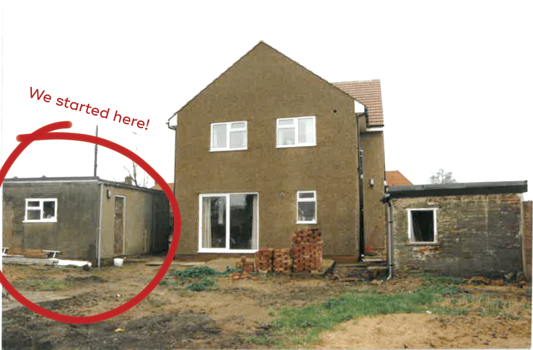 A view of a house in development, taken from a back garden. The house is in the middle of the photo, which looks old and taken on film. To the right is an outhouse of sorts, circled in red with the text 'we started here!'