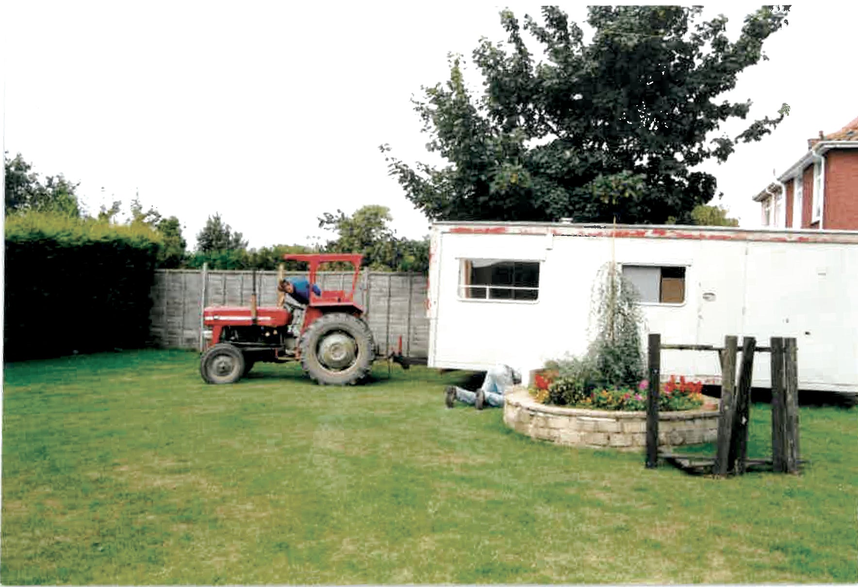 Taken from a back garden, the image looks like it is old and taken on film. It shows a small red tractor pulling a white caravan into the garden.