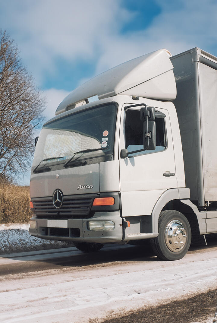 The front of a Mercedes Ateco lorry cab. Looks to be in summer sun, with a snowy and icy road, a bare tree in the background.